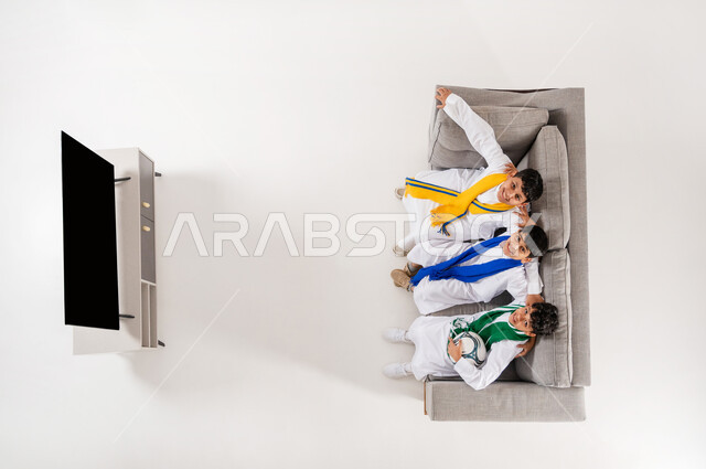 عرض شاشة سوداء فارغة, مشاركة الأصدقاء الأوقات الممتعة, بورتريه علوي عامودي لثلاثة أولاد عربيين سعوديين خليجيين يرتدون الثوب التقليدي يجلسون على الأريكة في غرفة المعيشة, متابعة مباراة كأس العالم, النظر الى الكاميرا بإيماءات التشجيع والحماس, خلفية بيضاء