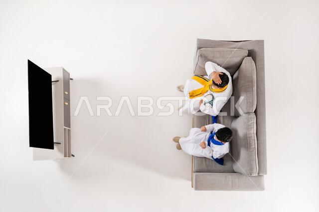 إيماءات التشجيع والحماس, عرض شاشة سوداء فارغة، مشاهدة مباريات كرة القدم, مشاركة الأصدقاء الأوقات الممتعة, بورتريه علوي عامودي لولدان عربيان خليجيان سعوديان يرتديان الثوب التقليدي يجلسان على الأريكة في غرفة المعيشة, متابعة مباراة كأس العالم، خلفية بيضاء