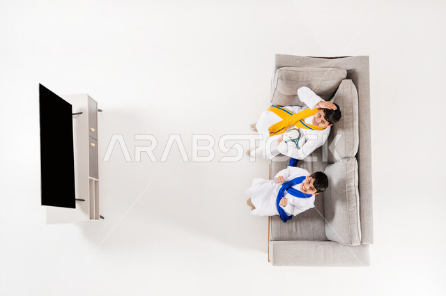 Watching football matches, sharing fun times with friends, vertical overhead portrait of two Saudi Arabian Gulf boys wearing traditional thobes sitting on sofa in living room, looking at camera with encouraging and enthusiastic gestures, showing blank black screen, white background