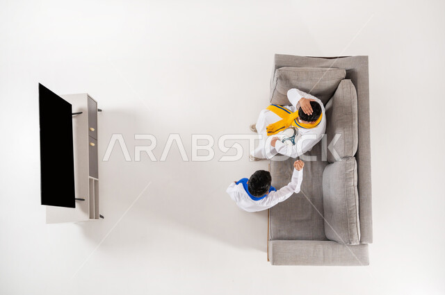 مشاركة الأصدقاء الأوقات الممتعة, بورتريه علوي عامودي لولدان عربيان خليجيان سعوديان يرتديان الثوب التقليدي يجلسان على الأريكة في غرفة المعيشة, متابعة مباراة كأس العالم، إيماءات التشجيع والحماس, عرض شاشة سوداء فارغة، مشاهدة مباريات كرة القدم, خلفية بيضاء