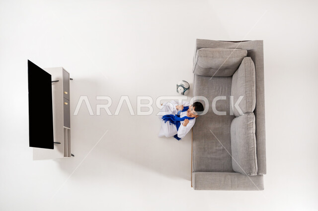 تشجيع المنتخب السعودي, عرض شاشة سوداء فارغة عبر التلفاز, مشاهدة مباريات كرة القدم, الشغف بالألعاب الرياضية , بورتريه علوي عامودي لولد عربي خليجي سعودي يرتدي الثوب التقليدي يجلس على الأرض في غرفة المعيشة , النظر إلى الكاميرا بتعابير التفاعل والحماس, خلفية بيضاء