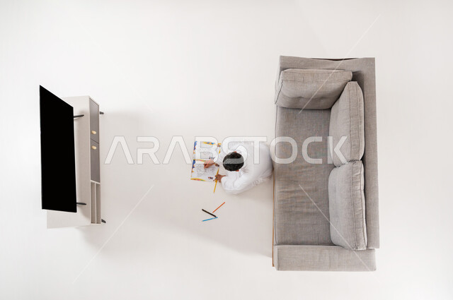 Practicing the hobby of drawing and coloring in free time, developing talents and doing fun recreational activities, displaying a blank black screen on TV, vertical portrait of a Saudi Arabian Gulf boy wearing traditional clothing sitting on the floor in the living room drawing on a notebook, white background