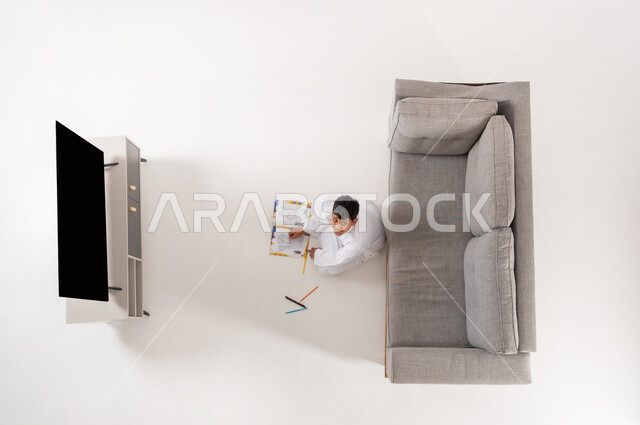 تنمية هواية الرسم و التلوين في أوقات الفراغ, ممارسة الأنشطة الترفيهية المنزلية, بورتريه علوي عامودي لولد عربي خليجي سعودي يرتدي الثوب التقليدي يجلس على الأرض في غرفة المعيشة, النظر إلى الكاميرا بإيماءات السرور والفرح, عرض شاشة سوداء فارغة, خلفية بيضاء