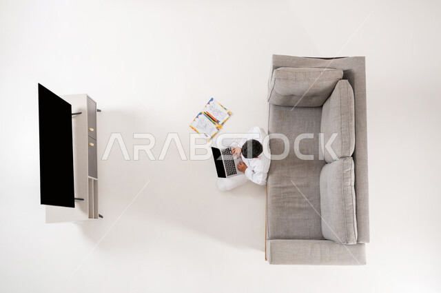 القيام بالأنشطة الترفيهية الممتعة, قضاء أوقات الفراغ بممارسة الهوايات, بورتريه علوي عامودي لولد عربي خليجي سعودي يرتدي الثوب التقليدي يجلس على الأرض في غرفة المعيشة يمسك جهاز التابلت بيديه, استخدام التكنولوجيا في الترفيه, عرض شاشة سوداء فارغة، خلفية بيضاء