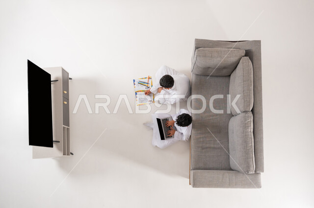 استغلال أوقات الفراغ بالأنشطة الالكترونية والواقعيه المفيدة، بورتريه علوي عامودي لأخوين عربيين خليجيين سعوديين يجلسان على الأرض في غرفة المعيشة, شاشة سوداء فارغة, قضاء وقت ممتع في تعلم الرسم والتلوين، إيماءات السعادة والفرح, خلفية بيضاء