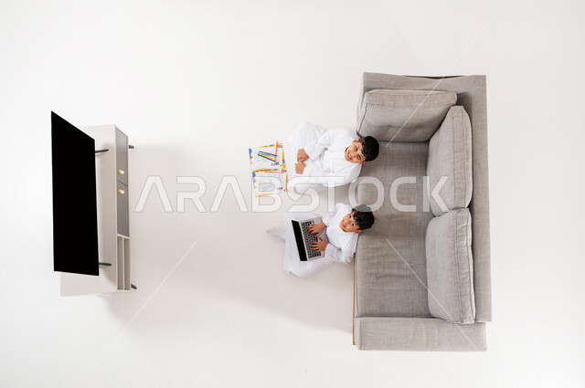 قضاء وقت ممتع في تعلم الرسم والتلوين، النظر الى الكاميرا بإيماءات السعادة والفرح, استغلال أوقات الفراغ بالأنشطة الالكترونية والواقعيه المفيدة، بورتريه علوي عامودي لأخوين عربيين خليجيين سعوديين يجلسان على الأرض في غرفة المعيشة, شاشة سوداء فارغة, خلفية بيضاء