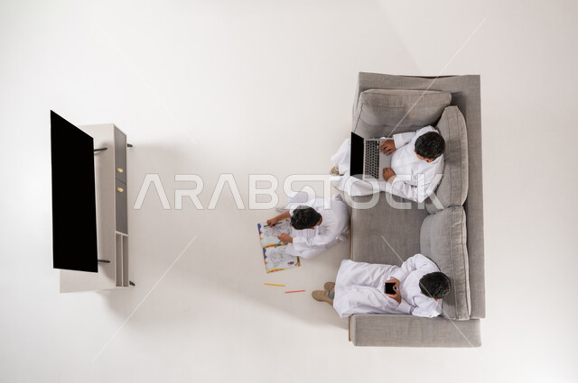 ممارسة الهوايات المسلية, استخدام التقنيات الحديثة في وقت الاستراحة, الترفيه والمتعة في أوقات الفراغ, بورتريه علوي عامودي لمجموعة أولاد عربيون خليجيون سعوديون يرتدون الثوب التقليدي السعودي يجلسون في غرفة المعيشة, الاستمتاع في هواية الرسم والتلوين, خلفية بيضاء