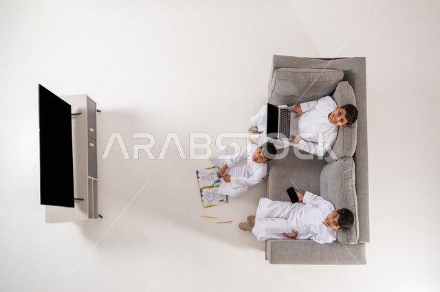 النظر إلى الكاميرا بايماءات السرور، استخدام التقنيات الحديثة في وقت الاستراحة, الترفيه والمتعة في أوقات الفراغ, بورتريه علوي عامودي  لاولاد عرب خليجيين سعوديين يرتدون الثوب التقليدي يجلسون في غرفة المعيشة يقومون بممارسة الهوايات المسلية, الاستمتاع في هواية الرسم والتلوين, خلفية بيضاء