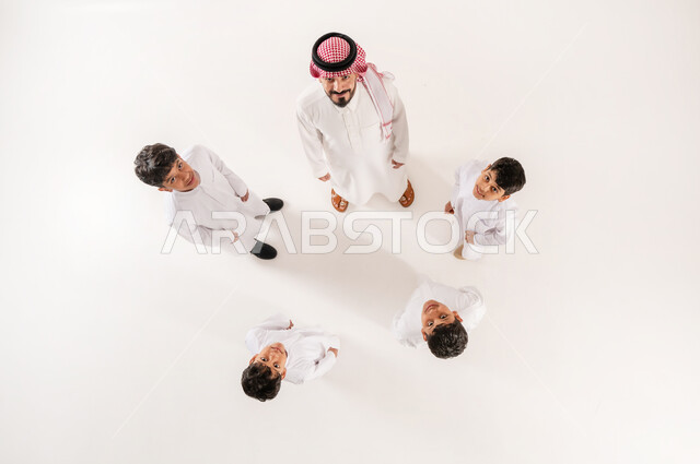 ايماءات السرور والسعادة، الالفة والانسجام والمودة بين افراد الاسرة، مساندة الأبناء و تشجيعهم، بورتريه علوي عامودي لرجل عربي خليجي سعودي يرتدي الثوب التقليدي والشماغ يقوم بقضاء وقت ممتع مع الاولاد، خلفية بيضاء