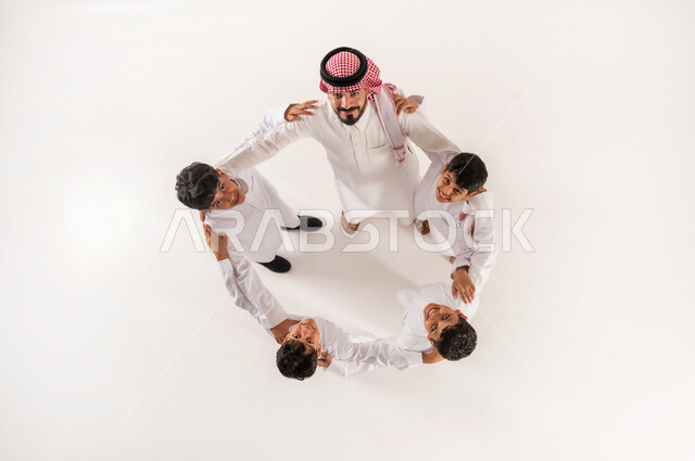 النظر الى الكاميرا بايماءات السرور والسعادة، مساندة الأبناء و تشجيعهم، الالفة والانسجام والمودة بين افراد الاسرة،  بورتريه علوي عامودي لرجل عربي خليجي سعودي يرتدي الثوب التقليدي والشماغ يضع يديه على أكتاف أولاده بتعابير الدعم والمحبة، خلفية بيضاء