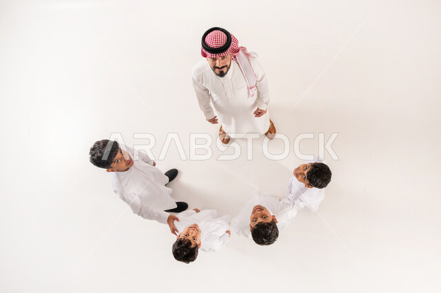 الالفة والانسجام والمودة بين افراد الاسرة،  قضاء وقت ممتع مع الاولاد، مساندة الأبناء و تشجيعهم، بورتريه علوي عامودي لرجل عربي خليجي سعودي يرتدي الثوب التقليدي والشماغ يقف برفقة اولاده ينظر الى الكاميرا بايماءات السرور والسعادة، خلفية بيضاء