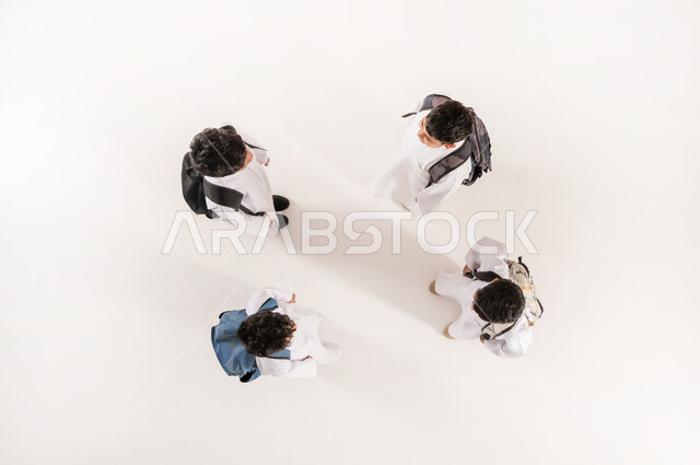 قضاء وقت ممتع مع الأصدقاء في المدرسة، الانتهاء من يوم دراسي، الألفة والانسجام و المودة، بورتريه علوي عامودي لمجموعة أولاد عرب خليجيين سعوديين يرتدون الثوب التقليدي يحملون حقائبهم يقفون برفقة بعضهم, خلفية بيضاء