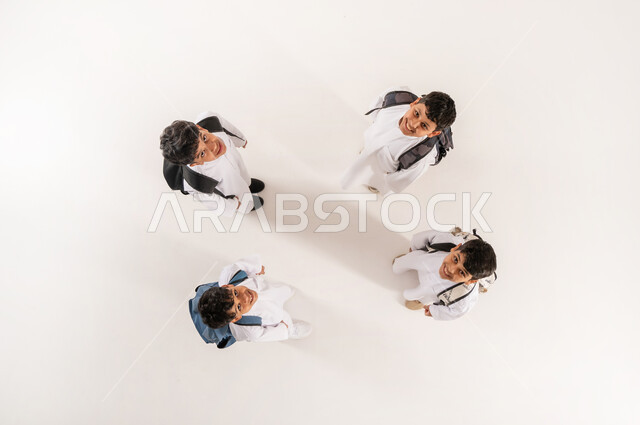 النظر إلى الگاميرا بايماءات السرور  والسعادة، قضاء وقت ممتع مع الأصدقاء في المدرسة، الانتهاء من يوم دراسي، الألفة والانسجام و المودة، بورتريه علوي عامودي لمجموعة أولاد عرب خليجيين سعوديين يرتدون الثوب التقليدي يحملون حقائبهم يقفون برفقة بعضهم, خلفية بيضاء