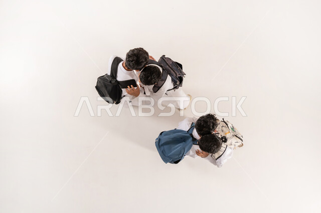 الألفة والانسجام و المودة، الانتهاء من اليوم الدراسي, مفهوم إلقاء التحية والوداع، قضاء وقت ممتع مع الأصدقاء في المدرسة، بورتريه علوي عامودي لمجموعة أولاد عرب خليجيين سعوديين يرتدون الثوب التقليدي يحملون حقائبهم يقومون باحتضان بعضهم البعض, خلفية بيضاء