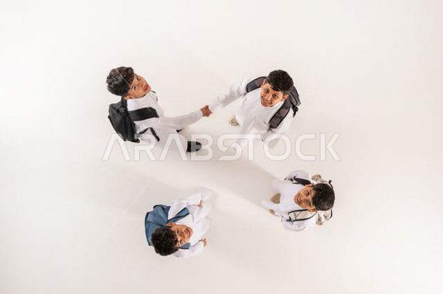 الانتهاء من اليوم الدراسي, مفهوم إلقاء التحية والوداع، قضاء وقت ممتع مع الأصدقاء في المدرسة، الألفة والانسجام و المودة، بورتريه علوي عامودي لمجموعة أولاد عرب خليجيين سعوديين يرتدون الثوب التقليدي يحملون حقائبهم يقومون بمصافحة بعضهم البعض, خلفية بيضاء