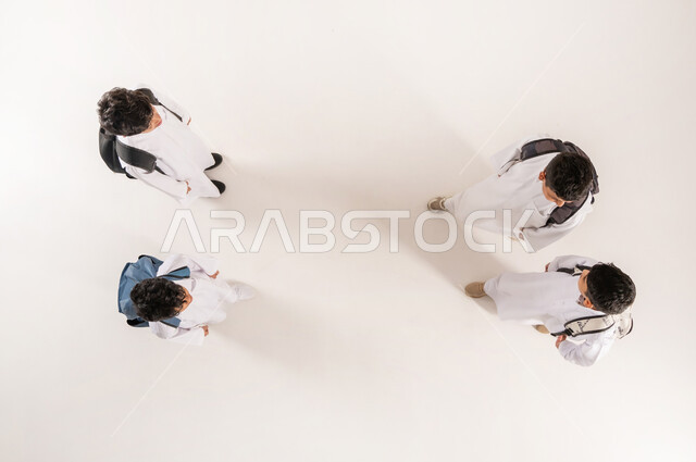 قضاء وقت ممتع مع الأصدقاء في المدرسة، الانتهاء من يوم دراسي، الألفة والانسجام و المودة، بورتريه علوي عامودي لمجموعة أولاد عرب خليجيين سعوديين يرتدون الثوب التقليدي يحملون حقائبهم يقفون برفقة بعضهم, خلفية بيضاء