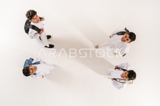 النظر إلى الگاميرا بايماءات السرور  والسعادة، قضاء وقت ممتع مع الأصدقاء في المدرسة، الانتهاء من يوم دراسي، الألفة والانسجام و المودة، بورتريه علوي عامودي لمجموعة أولاد عرب خليجيين سعوديين يرتدون الثوب التقليدي يحملون حقائبهم يقفون برفقة بعضهم, خلفية بيضاء