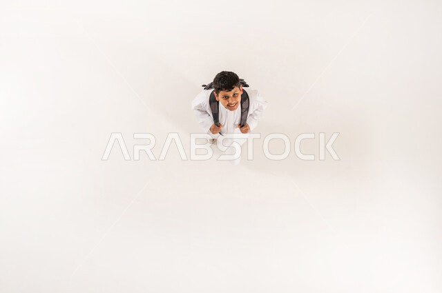 التحضر لعام دراسي جديد، العودة إلى الدراسة بهمة ونشاط، بورتريه علوي عامودي لصبي عربي خليجي سعودي يرتدي الثوب التقليدي يحمل حقيبة الظهر ينظر إلى الكاميرا بايماءات السرور, الاستعداد والأجهزة للذهاب الى المدرسة، خلفية بيضاء