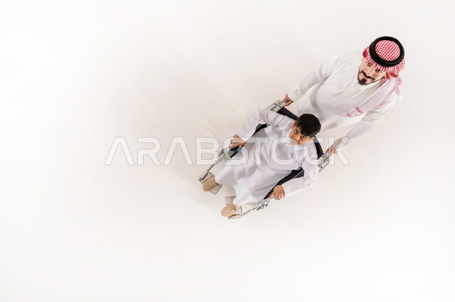 اهتمام ودعم المجتمع السعودي لذوي الاحتياجات الخاصة، تقديم الحب والرعاية لذوي الهمم، مساعدة مرضى الإعاقة الحركية على التنقل بسهولة, بورتريه  علوي عامودي لاب عربي خليجي سعودي يرتدي الثوب التقليدي الشماغ يقوم بمساعدة ولد المصاب بضعف القدرة الحركية على التنقل، العطف والحب بين أفراد الأسرة، خلفية بيضاء