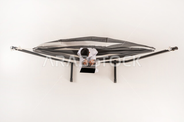 قضاء أوقات ممتعة في العطلة، دمج التقنيات الحديثة بالتسلية والمرح، مفهوم الاسترخاء والاستجمام، بورتريه علوي عامودي لولد عربي خليجي سعودي يرتدي الثوب التقليدي يجلس على أرجوحة قماشية معلقة يقوم بلعب العاب الفيديو باستخدام الحاسوب المحمول، خلفية بيضاء