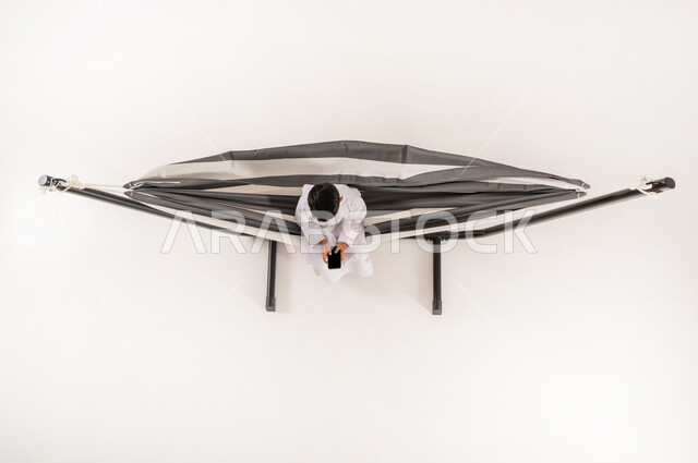 مفهوم الاسترخاء والاستجمام، قضاء أوقات ممتعة في العطلة، بورتريه علوي عامودي لولد عربي خليجي سعودي يرتدي الثوب التقليدي يجلس على أرجوحة قماشية معلقة يقوم بلعب العاب الفيديوباستخدام الهاتف المحمول، خلفية بيضاء