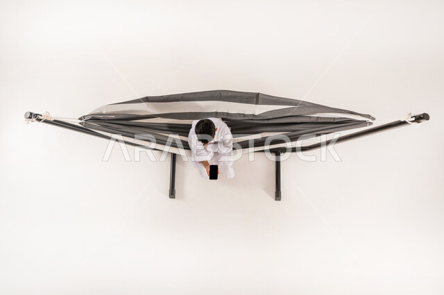 قضاء أوقات ممتعة في العطلة، مفهوم الاسترخاء والاستجمام، بورتريه علوي عامودي لولد عربي خليجي سعودي يرتدي الثوب التقليدي يجلس على أرجوحة قماشية معلقة يقوم باالعب العاب الفيديوباستخدام الهاتف المحمول، خلفية بيضاء