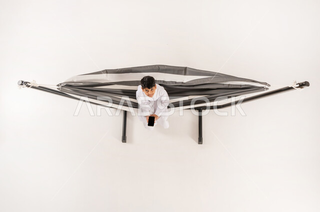 عرض شاشة سوداء فارغة، قضاء أوقات ممتعة في العطلة، مفهوم الاسترخاء والاستجمام، بورتريه علوي عامودي لولد عربي خليجي سعودي يرتدي الثوب التقليدي يجلس على أرجوحة قماشية معلقة يحمل بيده الهاتف المحمول ينظر الى الكاميرا بايماءات السرور، خلفية بيضاء