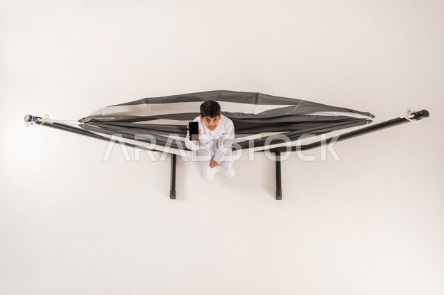 عرض شاشة سوداء فارغة، قضاء أوقات ممتعة في العطلة، مفهوم الاسترخاء والاستجمام، بورتريه علوي عامودي لولد عربي خليجي سعودي يرتدي الثوب التقليدي يجلس على أرجوحة قماشية معلقة يحمل بيده الهاتف المحمول ينظر الى الكاميرا بايماءات السرور، خلفية بيضاء