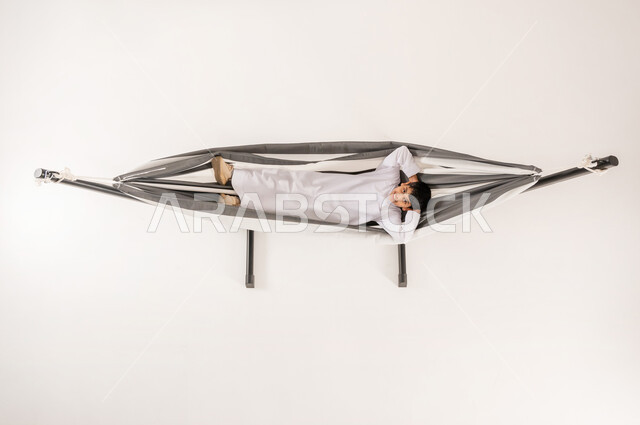 النظر إلى الكاميرا بتعابير البهجة والسرور، الحاجة إلى الراحة و الهدوء، مفهوم الاسترخاء والاستجمام، بورتريه علوي عامودي لولد عربي سعودي خليجي يرتدي الثوب التقليدي مستلقي على أرجوحة قماشية معلقة يضع يديه أسفل رأسه بايماءات الفرح والسعادة، خلفية بيضاء