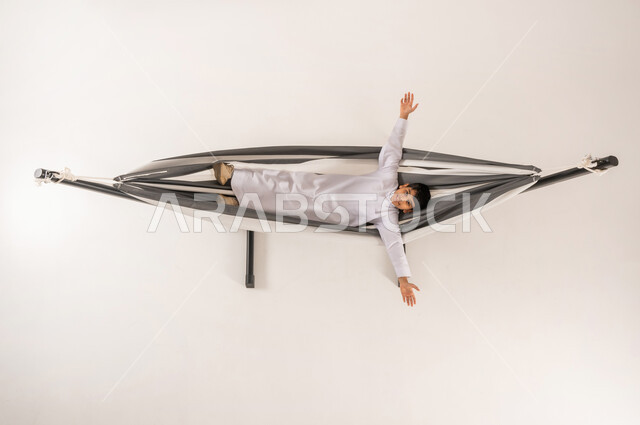 مفهوم الاسترخاء والاستجمام، بورتريه علوي عامودي لولد عربي سعودي خليجي يرتدي الثوب التقليدي يجلس على أرجوحة قماشية معلقة يرفع بايماءات المتعة والتسلية، تعابير البهجة والسرور، الحاجة إلى الراحة و الهدوء،خلفية بيضاء