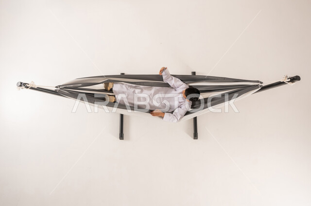 الاحتفاظ بالذكريات السعيدة، مفهوم الاسترخاء والاستجمام، بورتريه علوي عامودي لولد عربي خليجي سعودي يرتدي الثوب التقليدي يستلقي على أرجوحة قماشية معلقة يقوم بالتقاط صورة سيلفي باستخدام الهاتف المحمول، قضاء أوقات ممتعة في العطلة، خلفية بيضاء