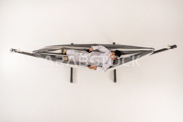 مفهوم الاسترخاء والاستجمام، دمج التقنيات الحديثة بالتسلية والمرح، قضاء أوقات ممتعة في العطلة، بورتريه علوي عامودي لولد عربي خليجي سعودي يرتدي الثوب التقليدي يستلقي على أرجوحة قماشية معلقة يحمل بيده الهاتف المحمول ينظر الى الكاميرا بايماءات السرور، خلفية بيضاء