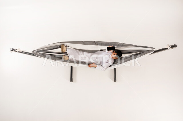 عرض شاشة سوداء فارغة، مفهوم الاسترخاء والاستجمام، قضاء أوقات ممتعة في العطلة، بورتريه علوي عامودي لولد عربي خليجي سعودي يرتدي الثوب التقليدي يستلقي على أرجوحة قماشية معلقة يحمل بيده الهاتف المحمول ينظر الى الكاميرا بايماءات السرور، خلفية بيضاء