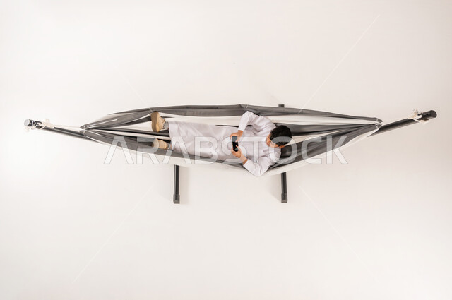 دمج التقنيات الحديثة بالتسلية والمرح، قضاء أوقات ممتعة في العطلة، مفهوم الاسترخاء والاستجمام، بورتريه علوي عامودي لولد عربي خليجي سعودي يرتدي الثوب التقليدي يستلقي على أرجوحة قماشية معلقة يقوم بلعب العاب الفيديو باستخدام الهاتف المحمول، خلفية بيضاء