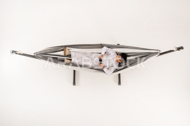 قضاء أوقات ممتعة في العطلة، دمج التقنيات الحديثة بالتسلية والمرح، مفهوم الاسترخاء والاستجمام، بورتريه علوي عامودي لولد عربي خليجي سعودي يرتدي الثوب التقليدي يستلقي على أرجوحة قماشية معلقة يقوم بلعب العاب الفيديو باستخدام الهاتف المحمول، خلفية بيضاء