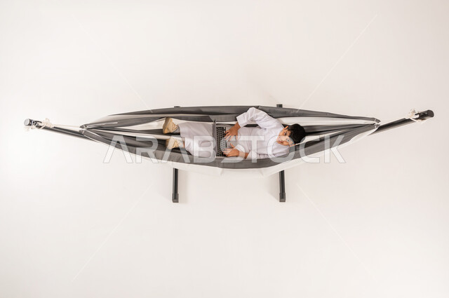 دمج التقنيات الحديثة بالتسلية والمرح، مفهوم الاسترخاء والاستجمام، قضاء أوقات ممتعة في العطلة، بورتريه علوي عامودي لولد عربي خليجي سعودي يرتدي الثوب التقليدي يجلس على أرجوحة قماشية معلقة يقوم بلعب العاب الفيديو باستخدام الحاسوب المحمول، خلفية بيضاء