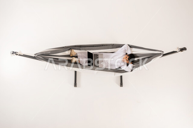 مفهوم الاسترخاء والاستجمام، قضاء أوقات ممتعة في العطلة، مشاهدة افلام الكارتون المسلية، دمج التقنيات الحديثة بالتسلية واللعب، بورتريه علوي عامودي لولد عربي خليجي سعودي يرتدي الثوب التقليدي يجلس على أرجوحة قماشية معلقة يقوم باستخدام الحاسوب المحمول، خلفية بيضاء