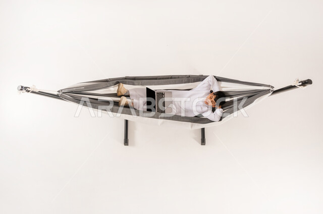 قضاء أوقات ممتعة في العطلة، مشاهدة افلام الكارتون المسلية، مفهوم الاسترخاء والاستجمام، دمج التقنيات الحديثة بالتسلية واللعب، بورتريه علوي عامودي لولد عربي خليجي سعودي يرتدي الثوب التقليدي يجلس على أرجوحة قماشية معلقة يقوم باستخدام الحاسوب المحمول، خلفية بيضاء