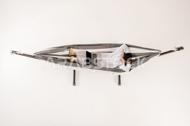 مشاهدة افلام الكارتون المسلية، قضاء أوقات ممتعة في العطلة، مفهوم الاسترخاء والاستجمام، دمج التقنيات الحديثة بالتسلية واللعب، بورتريه علوي عامودي لولد عربي خليجي سعودي يرتدي الثوب التقليدي يجلس على أرجوحة قماشية معلقة يقوم باستخدام الحاسوب المحمول، خلفية بيضاء