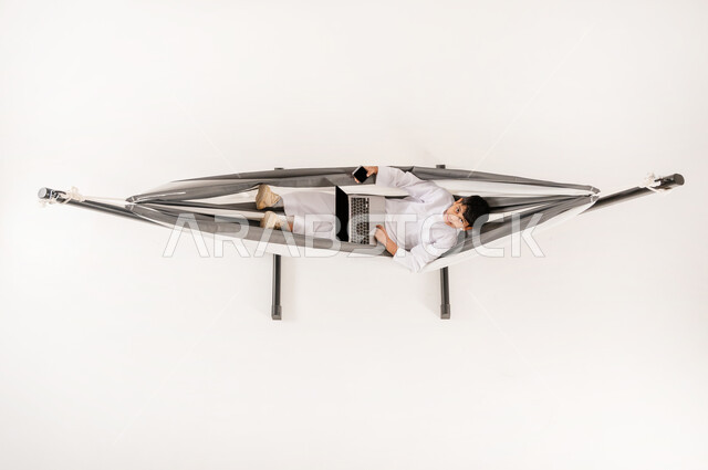 مفهوم الاسترخاء والاستجمام، قضاء أوقات ممتعة في العطلة، مشاهدة افلام الكارتون المسلية، دمج التقنيات الحديثة بالتسلية واللعب، بورتريه علوي عامودي لولد عربي خليجي سعودي يرتدي الثوب التقليدي يجلس على أرجوحة قماشية معلقة يحمل الهاتف والحاسوب المحمول، خلفية بيضاء