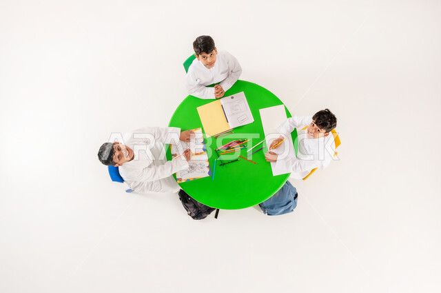النظر الى الكاميرا بايماءات السرور والسعادة، تعلم مهارات التركيز والابتكار، مفهوم الاهتمام بالهوايات، تعزيز المواهب الابداعية عند الطلاب، أنشطة ترفيهية مسلية، بورتريه علوي عامودي لتلاميذ عرب خليجيين سعوديين يرتدون الزي التقليدي يقومون بالرسم والتلوين، خلفية بيضاء