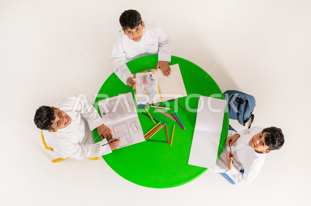 النظر الى الكاميرا بايماءات السرور والسعادة، أنشطة ترفيهية مسلية، بورتريه علوي عامودي لتلاميذ عرب خليجيين سعوديين يرتدون الزي التقليدي يقومون بالرسم والتلوين، تعزيز المواهب الابداعية عند الطلاب، مفهوم الاهتمام بالهوايات، تعلم مهارات التركيز والابتكار، خلفية بيضاء