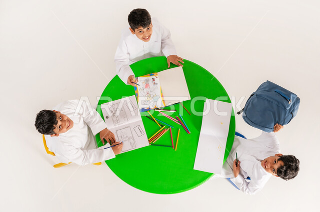 أنشطة ترفيهية مسلية، بورتريه علوي عامودي لتلاميذ عرب خليجيين سعوديين يرتدون الزي التقليدي يقومون بالرسم والتلوين، النظر الى الكاميرا بايماءات السرور والسعادة، تعزيز المواهب الابداعية عند الطلاب، مفهوم الاهتمام بالهوايات، تعلم مهارات التركيز والابتكار، خلفية بيضاء