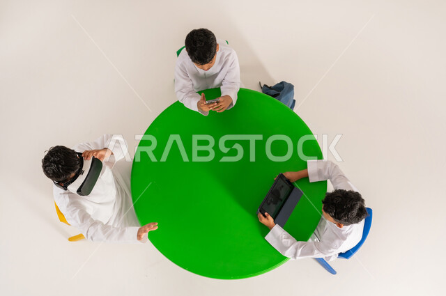 مفهوم محاكاة العالم الخيالي، دمج الواقع الافتراضي بالتعلم والتدريب, بورتريه علوي عامودي لطلاب عرب خليجيين سعوديين يرتدون الثوب التقليدي يقومون باستخدام التقنيات الحديثة والمتطورة في التعلم, خلفية بيضاء