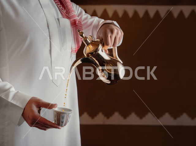 مفهوم الكرم وحسن الاستقبال، ذكرى تأسيس الدولة السعودية الاولى 22 فبراير، يوم بدينا 1727 م، الاحتفال بالمناسبات والأعياد الوطنية، صورة مقربة لرجل عربي خليجي سعودي يرتدي الأزياء التراثية يقوم بصب القهوة من الدلة النحاسية، تقديم المنتجات المحلية للضيوف