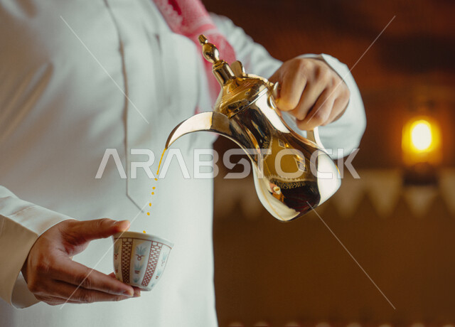 مفهوم الكرم وحسن الاستقبال، ذكرى تأسيس الدولة السعودية الاولى 22 فبراير، يوم بدينا 1727 م، الاحتفال بالمناسبات والأعياد الوطنية، صورة مقربة لرجل عربي خليجي سعودي يرتدي الأزياء التراثية يقوم بصب القهوة من الدلة النحاسية، تقديم المنتجات المحلية للضيوف