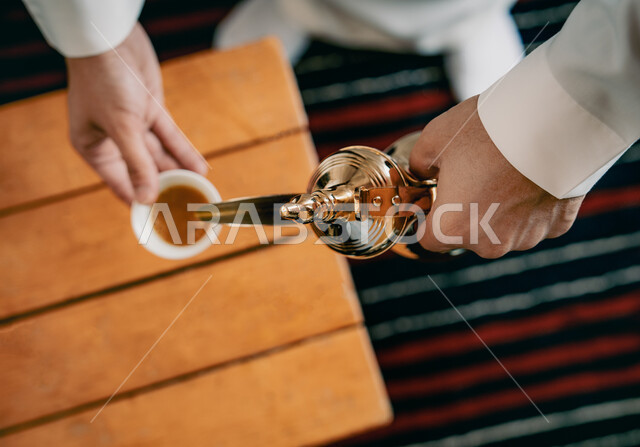 تقديم المنتجات المحلية للضيوف، مفهوم الكرم وحسن الاستقبال، ذكرى تأسيس الدولة السعودية الاولى 22 فبراير، يوم بدينا 1727 م، الاحتفال بالمناسبات والأعياد الوطنية، صورة مقربة لرجل عربي خليجي سعودي يرتدي الأزياء التراثية يقوم بصب القهوة من الدلة النحاسية 