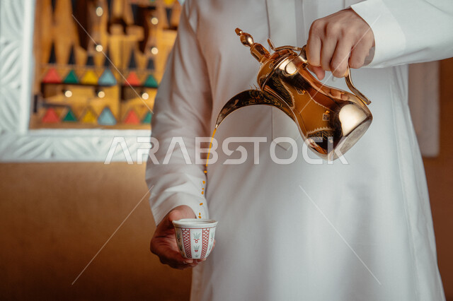 تقديم المنتجات المحلية للضيوف، مفهوم الكرم وحسن الاستقبال، ذكرى تأسيس الدولة السعودية الاولى 22 فبراير، يوم بدينا 1727 م، الاحتفال بالمناسبات والأعياد الوطنية، صورة مقربة لرجل عربي خليجي سعودي يرتدي الأزياء التراثية يقوم بصب القهوة من الدلة النحاسية 
