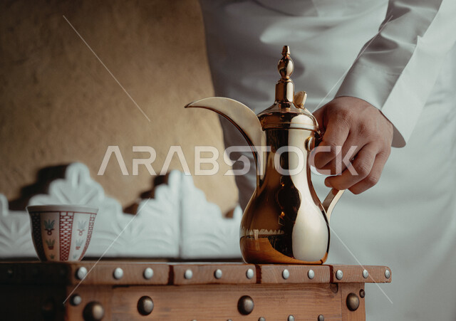 تقديم المنتجات المحلية للضيوف، مفهوم الكرم وحسن الاستقبال، ذكرى تأسيس الدولة السعودية الاولى 22 فبراير، يوم بدينا 1727 م، الاحتفال بالمناسبات والأعياد الوطنية، صورة مقربة لرجل عربي خليجي سعودي يرتدي الأزياء التراثية يمسك بيده دلة نحاسية لصب القهوة