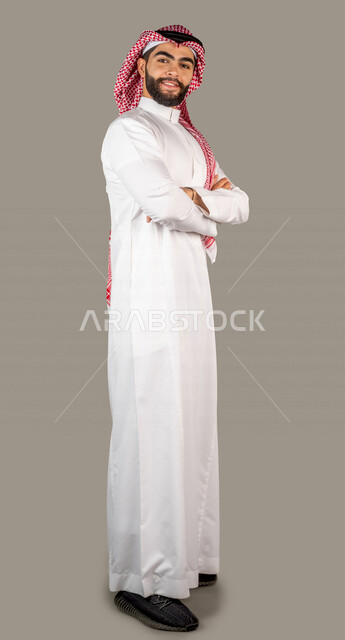 بورتريه لرجل عربي خليجي سعودي وسيم مكتف يديه يرتدي الزي السعودي التقليدي ، ايماءات و تعابير للوجه و اليدين ، خلفية رمادية ، كامل الجسم ، صورة بالطول الكامل